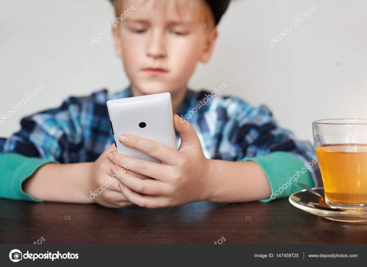 A little schoolboy dressed in checked shirt siting at wooden table with a cup of tea holding smartphone in his hands. People, technology, lifestyle and internet concept